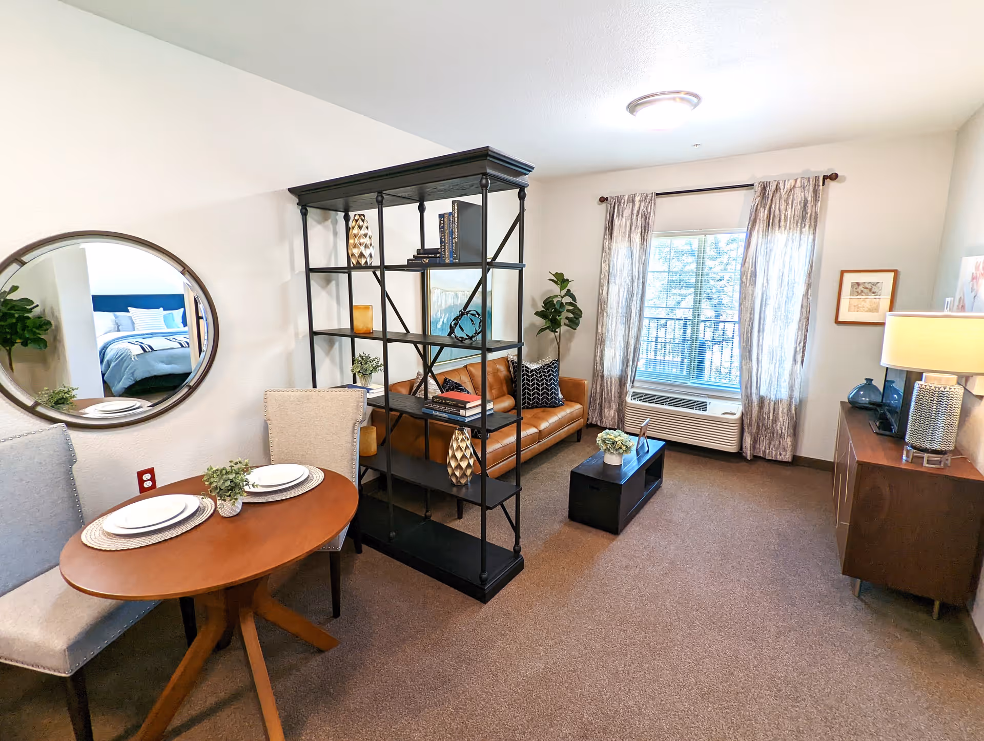 A cozy living room area with a brown leather sofa, a black metal and wood shelving unit, a small wooden coffee table with a plant, and a wooden sideboard with a lamp and decorative items. There is a window with patterned curtains letting in natural light. A round mirror on the wall reflects a bedroom with a bed and pillows. A small dining table with two upholstered chairs and place settings is also visible.
