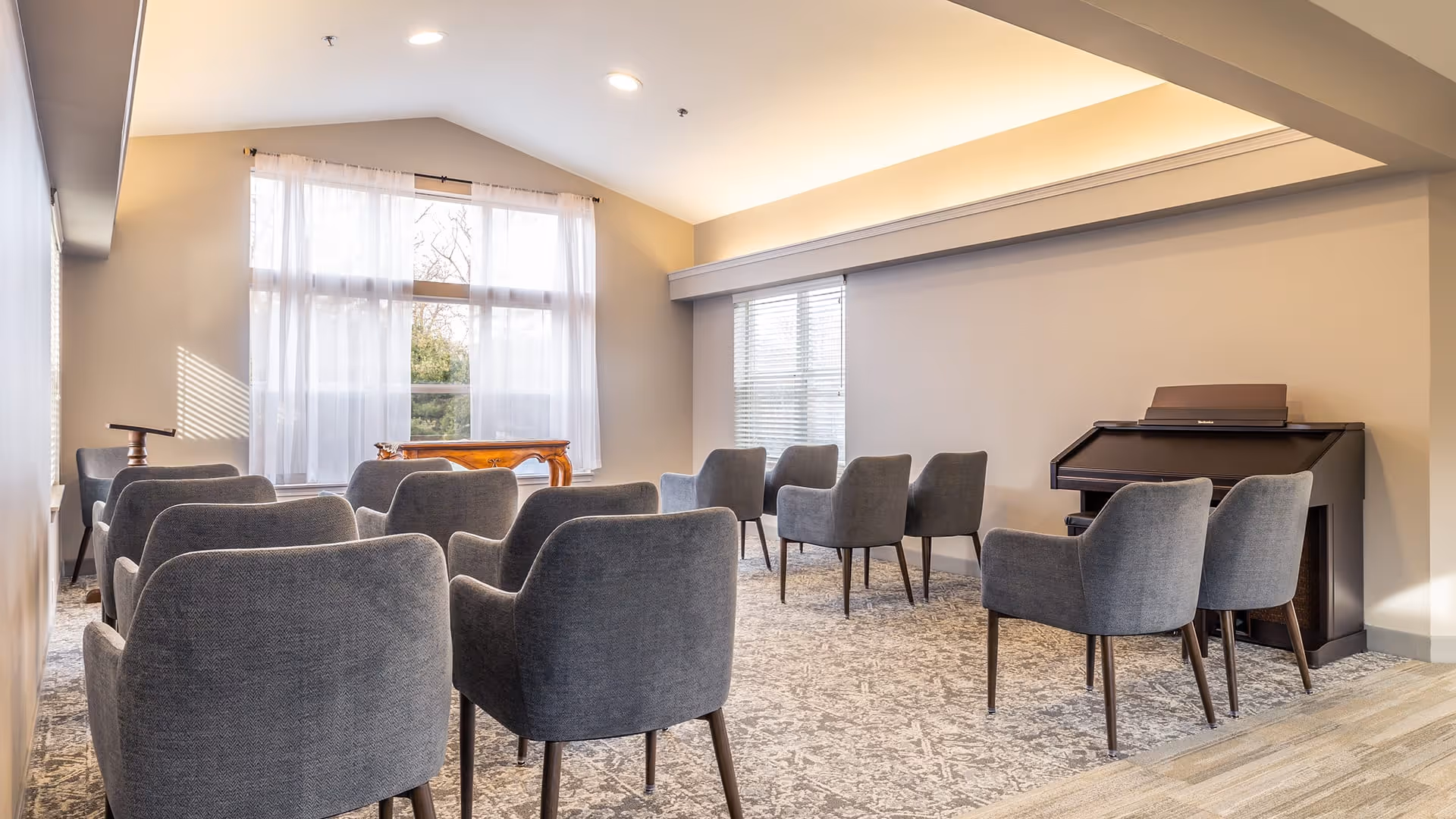 A bright room with multiple gray upholstered chairs arranged in rows facing a wooden piano against the wall. Large windows with sheer white curtains allow natural light to fill the space, and the ceiling has recessed lighting and a tray design.
