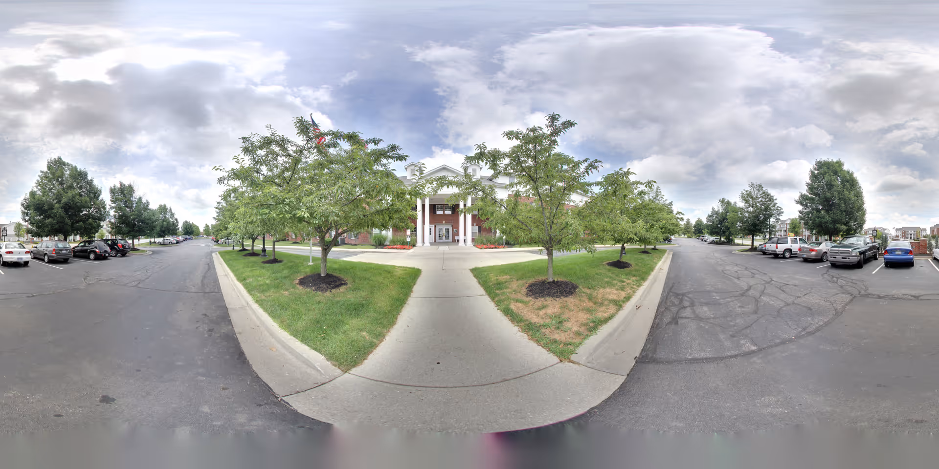 Wide view of the front exterior of a senior living facility named Benjamin Court, showing a paved driveway and parking areas on both sides with several parked cars. The building has a brick facade with white columns and a covered entrance. Trees and green grass line the walkway leading to the entrance under a partly cloudy sky.