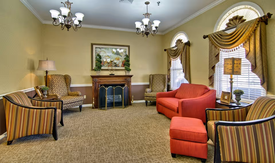 A cozy living room with a red sofa and matching ottoman, two striped armchairs, and two patterned wingback chairs arranged around a wooden fireplace with decorative plants and a framed picture above it. The room has beige walls with a brown wainscot, two large arched windows with ornate drapes, and two ceiling chandeliers providing light.