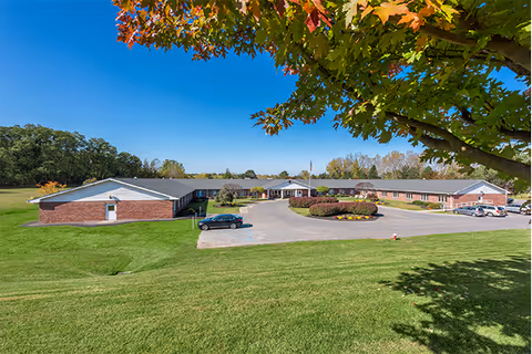 Front exterior of a single-story brick senior living facility with a circular driveway, flagpole, and landscaped lawn under a clear blue sky.