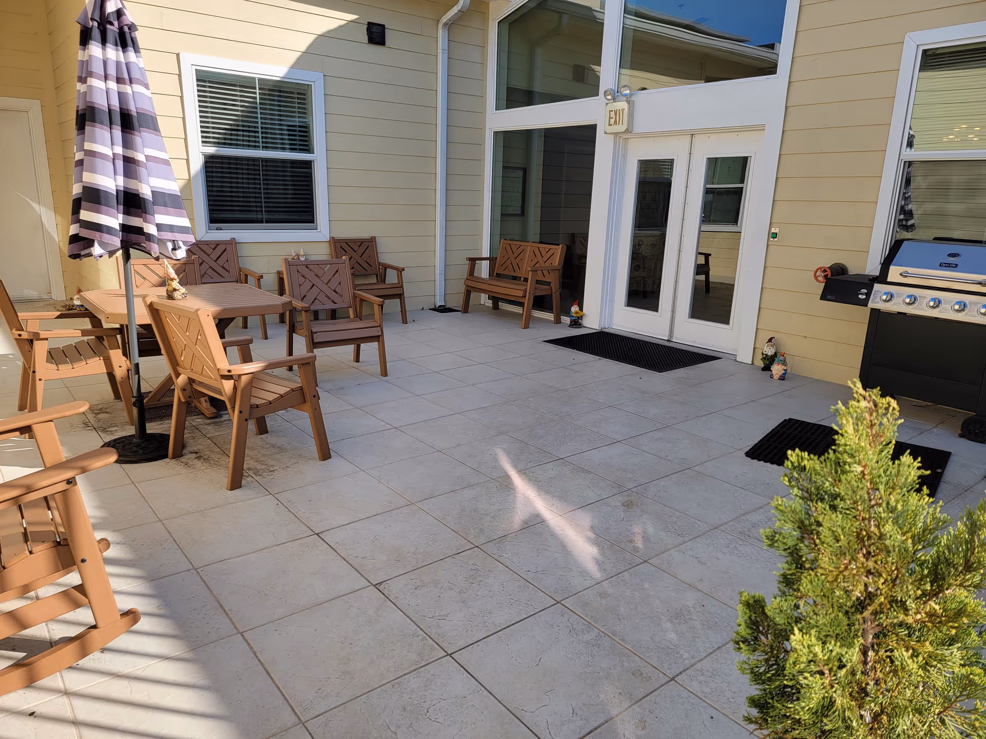Outdoor patio area with beige tiled flooring, several brown wooden chairs and benches, a table with a striped umbrella, a barbecue grill, and two small garden gnome statues near a set of white double doors with an exit sign above them. A small green shrub is visible in the foreground.
