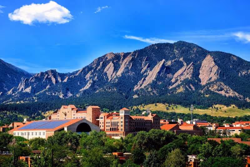 Red‑roofed brick buildings and greenery set beneath a dramatic mountain range under a bright blue sky.