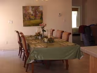 A dining room with a long table covered by a green tablecloth, place settings and floral centerpieces, surrounded by chairs and a painting on the wall.