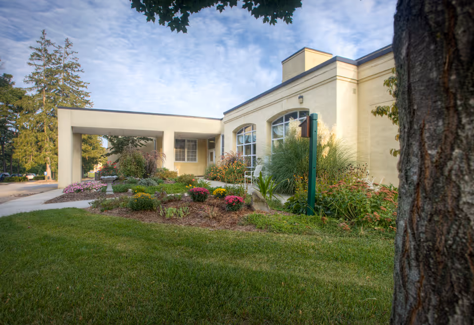 Exterior view of a single-story building with beige walls and large windows, surrounded by a well-maintained garden with colorful flowers and green shrubs. A tree trunk is visible in the foreground on the right side, and a covered walkway extends from the building entrance. The sky is partly cloudy.