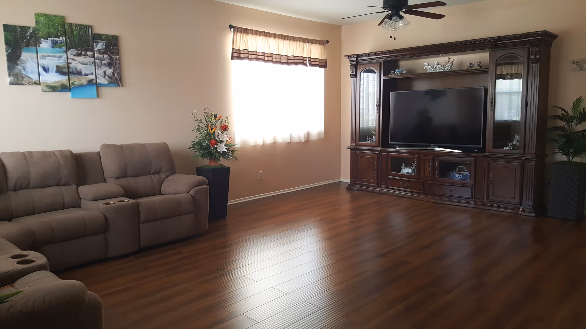A living room with wooden flooring, a large brown entertainment center with a flat-screen TV, a ceiling fan with lights, a brown reclining sofa with cup holders, a window with sheer curtains, two potted plants, and a multi-panel wall art depicting a waterfall scene.