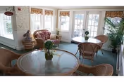 Sunlit sitting room with wicker tables and chairs, potted plants, and large windows and French doors.