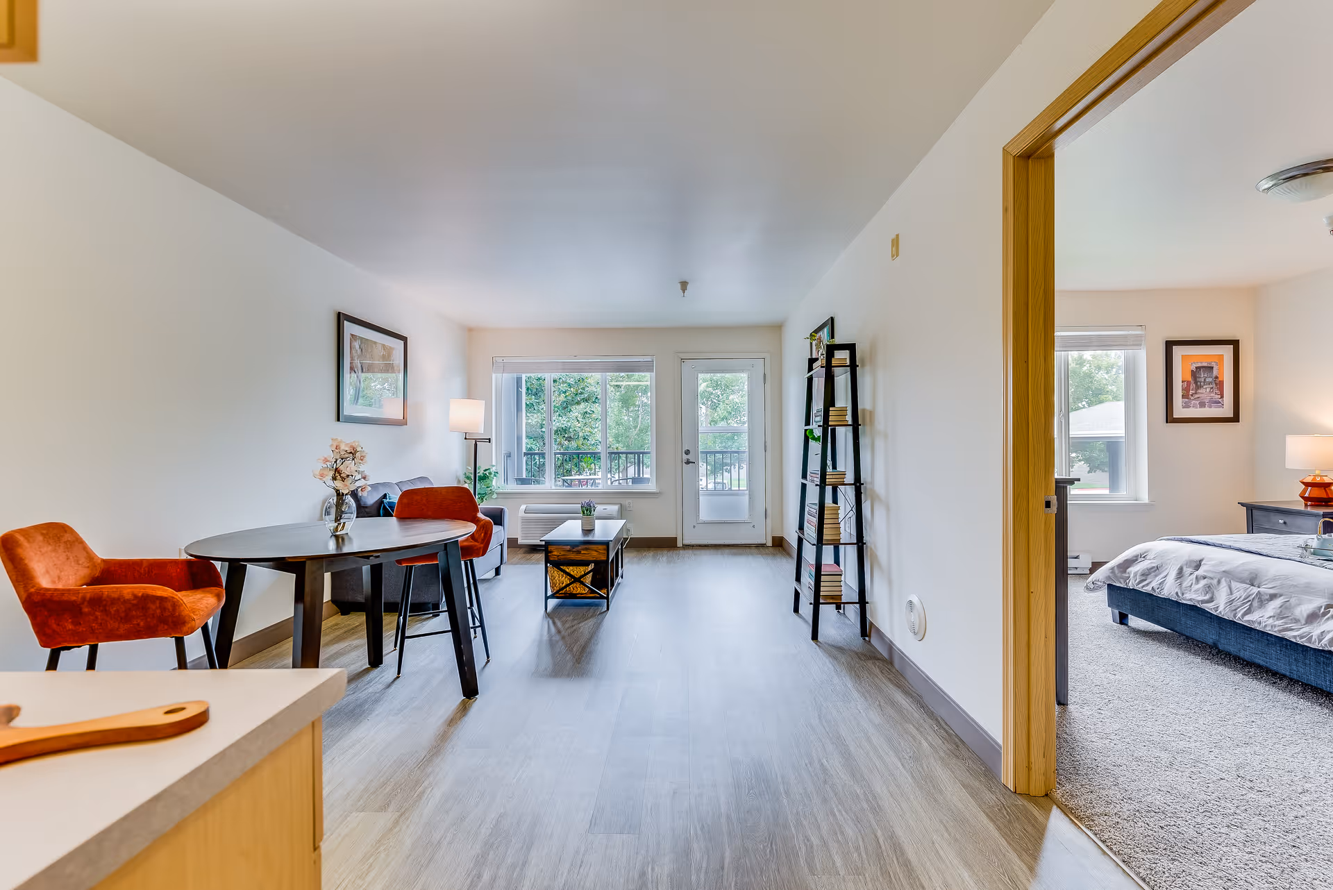 Bright and spacious senior living apartment interior featuring a small dining area with two orange chairs and a round table, a living area with a sofa, coffee table, and a tall bookshelf, and a bedroom visible through an open doorway with a bed, nightstand, and window. Large windows and a glass door provide natural light and a view of greenery outside.
