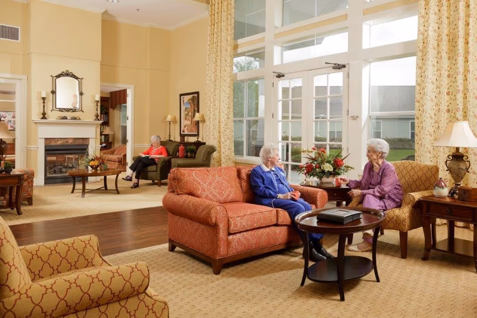 A bright and spacious living room in an assisted living facility with large windows and glass doors letting in natural light. The room is furnished with comfortable armchairs and sofas in warm tones, a coffee table with a floral arrangement, and a fireplace with a mirror and candles above it. Three elderly women are seated, two engaged in conversation and one reading a book in the background.