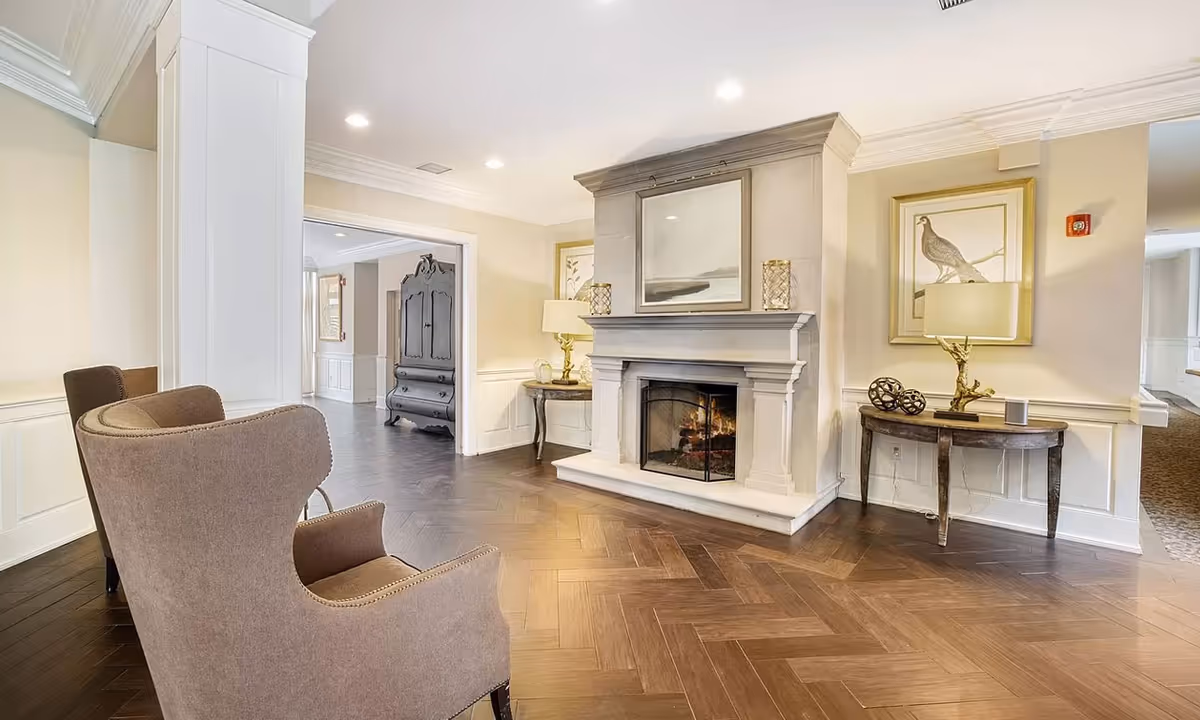 Bright seating area with a lit fireplace, two wingback chairs, herringbone wood floor, and decorative tables with lamps.