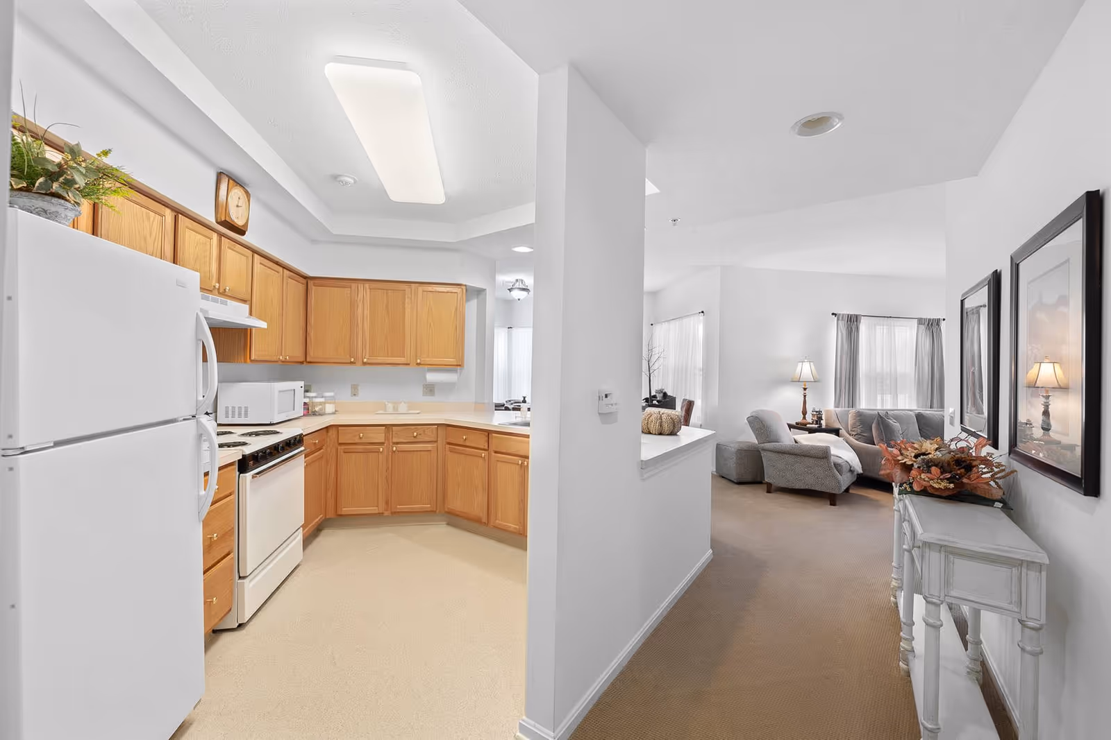 Interior view of a senior living facility showing a kitchen with wooden cabinets, white refrigerator, stove, and microwave on the left side, and a living room area with a sofa, armchair, side table with a lamp, and framed pictures on the right side. The walls are white and the space is well-lit with natural light coming through the windows.