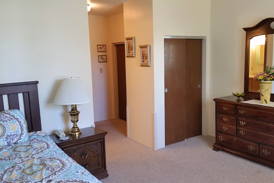 A bedroom with a wooden bed frame partially visible on the left, a nightstand with a brass lamp and a telephone, beige carpet, a closet with wooden sliding doors, a wooden dresser with a mirror, and framed pictures on the walls.