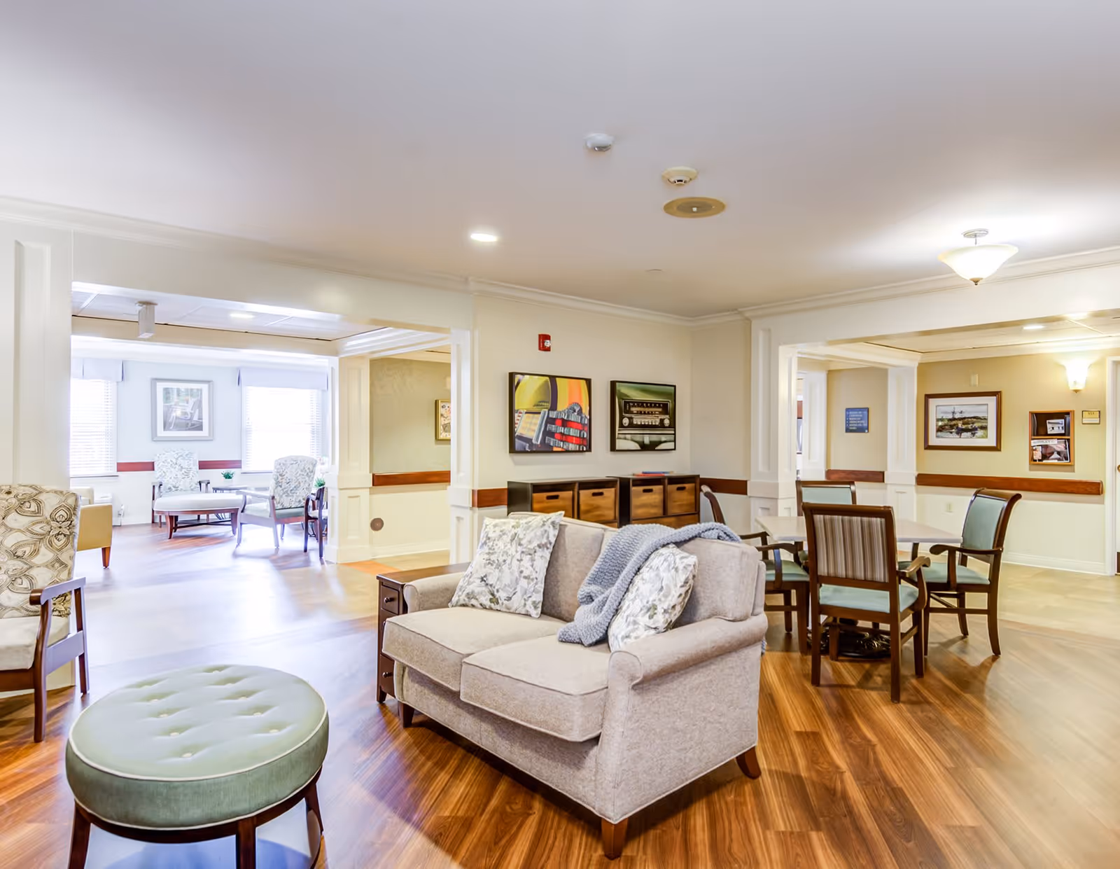 A bright and spacious senior living common area with wooden flooring, a beige loveseat with patterned pillows and a gray throw blanket, a round green ottoman, and several wooden chairs and tables. The walls are decorated with framed artwork, and large windows allow natural light to fill the space.