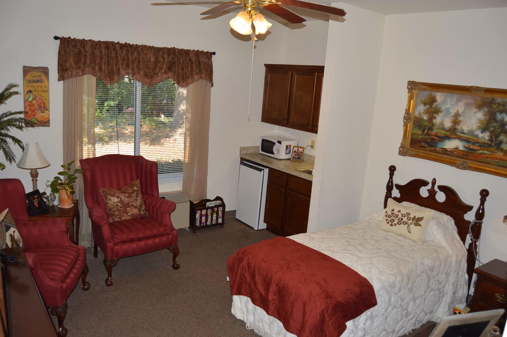 A cozy senior living room with a single bed covered in a white quilt and a red throw blanket. There are two red upholstered armchairs with decorative pillows near a window with beige curtains and a valance. A small kitchenette area with wooden cabinets, a microwave, and a mini fridge is visible. The room is decorated with a framed landscape painting above the bed and a wall hanging with a Thanksgiving theme. A ceiling fan with lights is mounted on the ceiling.
