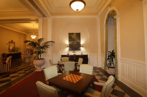 Warmly lit common room featuring a central game table with a chessboard surrounded by upholstered chairs, decorative molding, plants, and a sideboard.