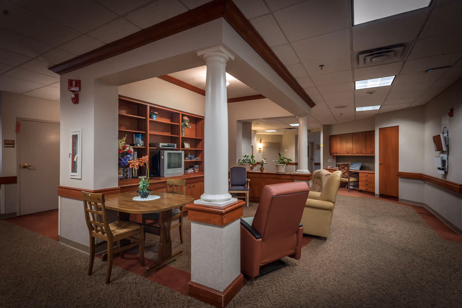 Interior view of a senior living facility common area with a small round wooden table and two chairs, a built-in wooden bookshelf with a TV and decorative items, several upholstered armchairs, potted plants, and a desk with a chair in the background. The space features white columns, beige walls, and a carpeted floor under fluorescent ceiling lights.
