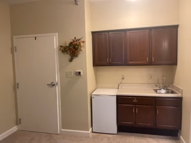 Small kitchenette area with dark wooden cabinets above and below a countertop. There is a small white refrigerator under the counter and a stainless steel sink on the right side. To the left of the kitchenette is a closed white door and a wall decorated with a small autumn-themed wreath.