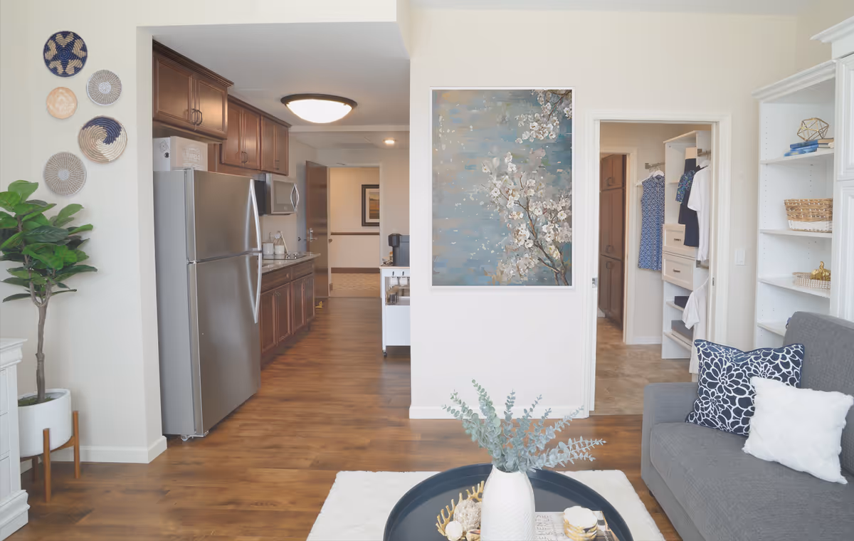 Interior view of a senior living facility showing a kitchen with stainless steel refrigerator and wooden cabinets on the left, a living area with a gray couch and decorative pillows on the right, a coffee table with a white vase and greenery in the center, and a hallway leading to a closet with hanging clothes. The walls are decorated with a painting of white flowers and several woven baskets.