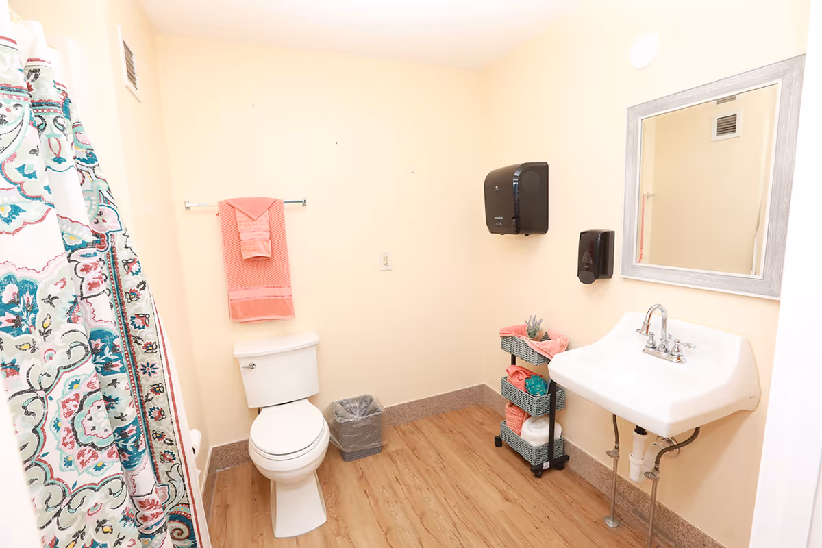 A clean bathroom with a white toilet, a white sink with a silver faucet, a mirror above the sink, a colorful patterned shower curtain, a towel rack with pink towels, a small trash bin, and a three-tiered storage cart holding towels and other items. The walls are painted light beige and the floor has a wood-like finish.