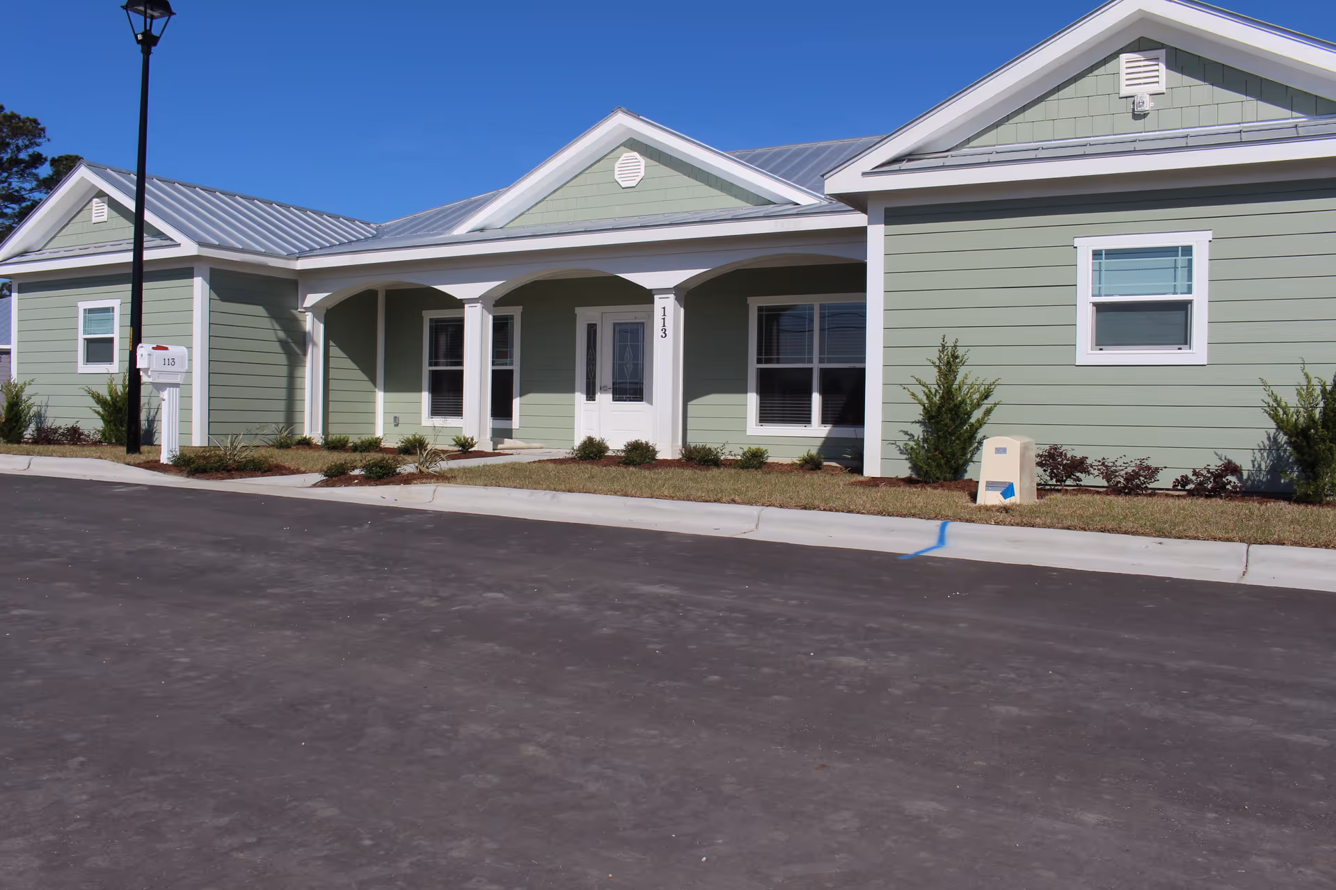 Exterior view of a single-story green building with white trim and a metal roof under a clear blue sky. The building has a covered porch with three arches and a white front door with sidelights. There are small shrubs and plants along the front of the building, a street lamp, and a mailbox labeled 113 near the curb.