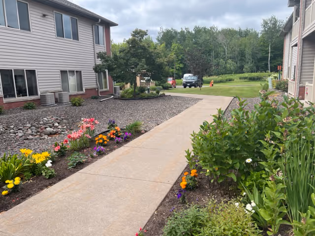 A paved walkway between two buildings with flower beds on both sides containing colorful flowers and green shrubs. In the background, there are trees and a parked vehicle under a cloudy sky.
