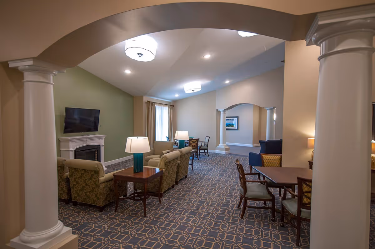 Interior view of a senior living facility lounge area with patterned carpet, beige walls, and white columns. The room features multiple seating arrangements including armchairs and tables with lamps. A flat-screen TV is mounted above a white fireplace on a green accent wall. There are windows with curtains allowing natural light into the space.