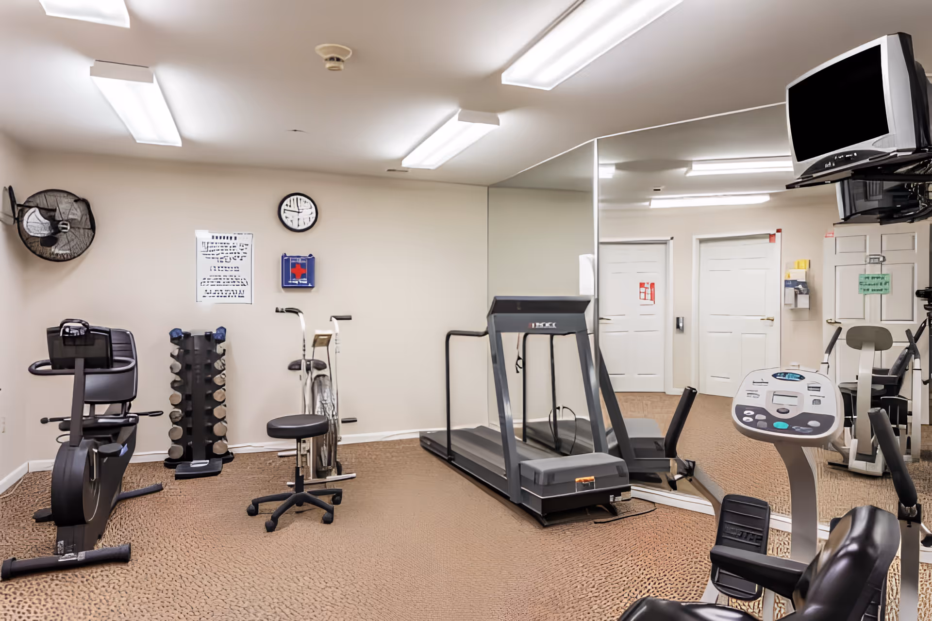 A small fitness room with exercise equipment including a treadmill, stationary bike, weight rack, and other cardio machines. The room has beige walls, a large mirror on one side, fluorescent ceiling lights, a wall clock, and a mounted TV in the corner.