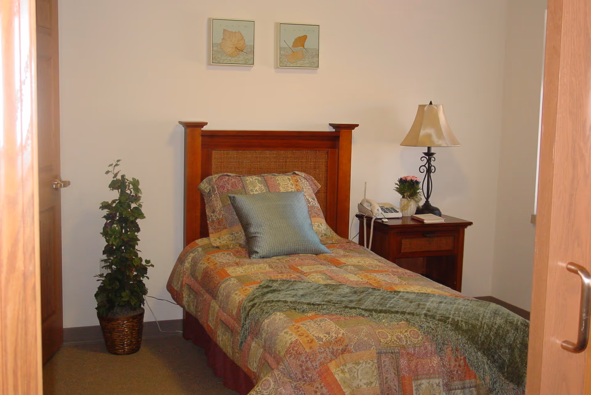 A small bedroom with a single bed covered in a multicolored patchwork quilt and a green throw blanket. There is a wooden headboard, a blue decorative pillow, and two framed leaf prints on the wall above the bed. To the right of the bed is a wooden nightstand with a lamp, a telephone, a small plant, and a book. A potted plant is placed on the floor to the left of the bed. The room has beige walls and carpeted floor.