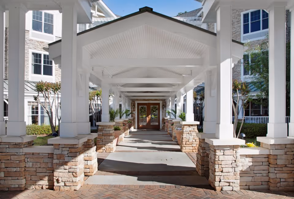Covered walkway entrance to a senior living facility with white pillars and stone bases, leading to double wooden doors. The building exterior features stone and white siding with multiple windows and landscaping including small trees and shrubs.