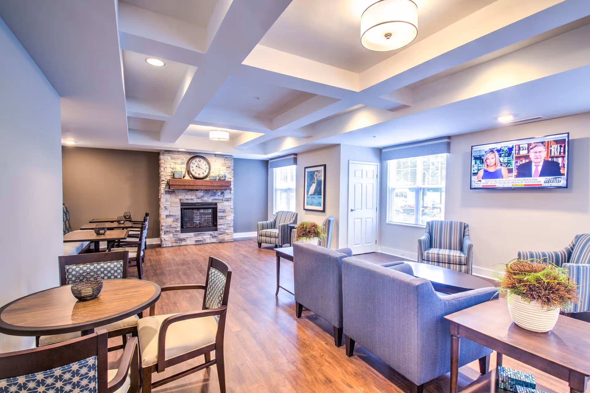 A bright and spacious senior living common area with wooden flooring, a stone fireplace with a clock above it, multiple seating arrangements including blue upholstered chairs and sofas, round wooden tables with chairs, a wall-mounted TV showing a news program, and large windows allowing natural light to fill the room.