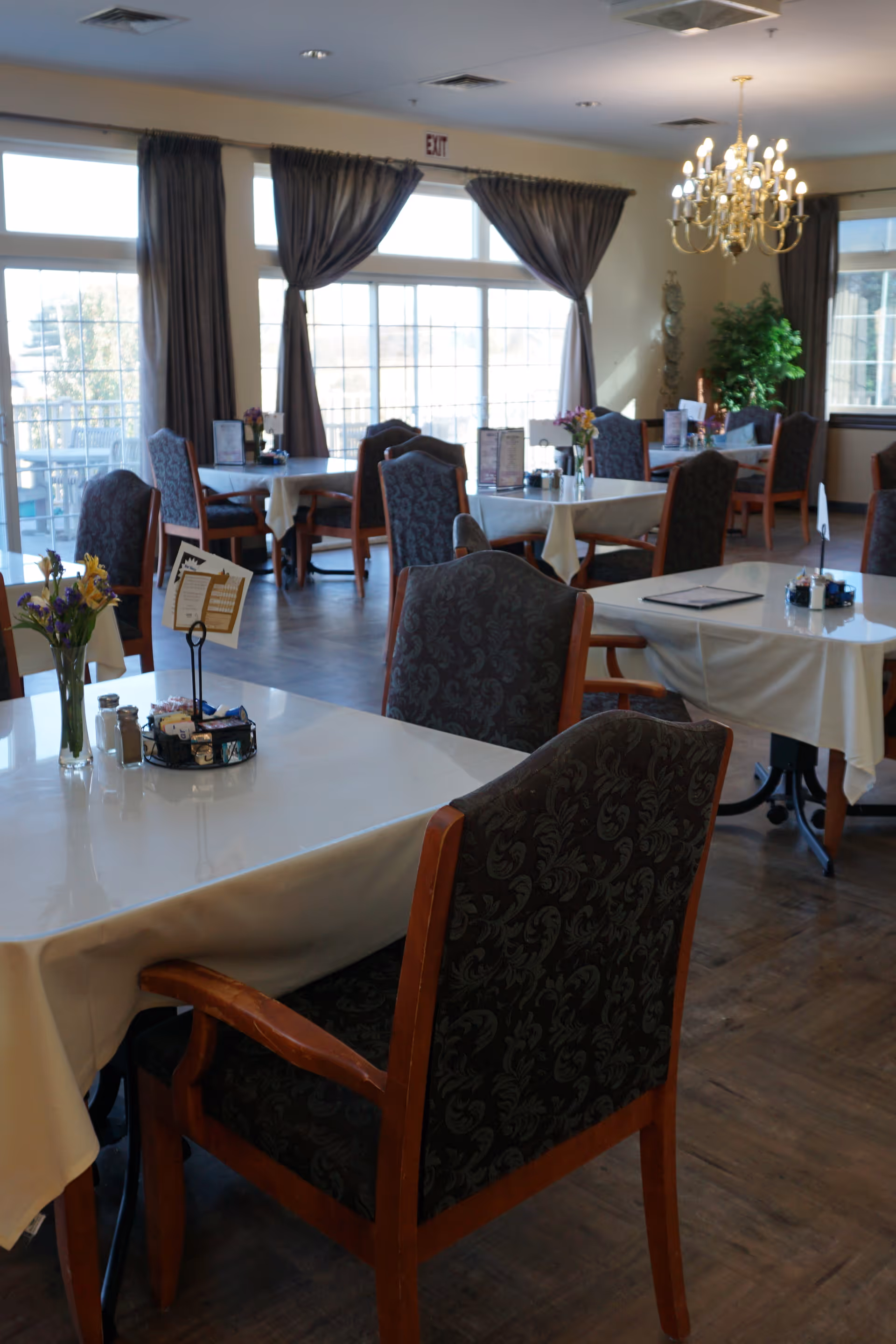 A bright dining room with several tables covered in white cloths, upholstered wooden chairs, large windows with curtains, and a chandelier.