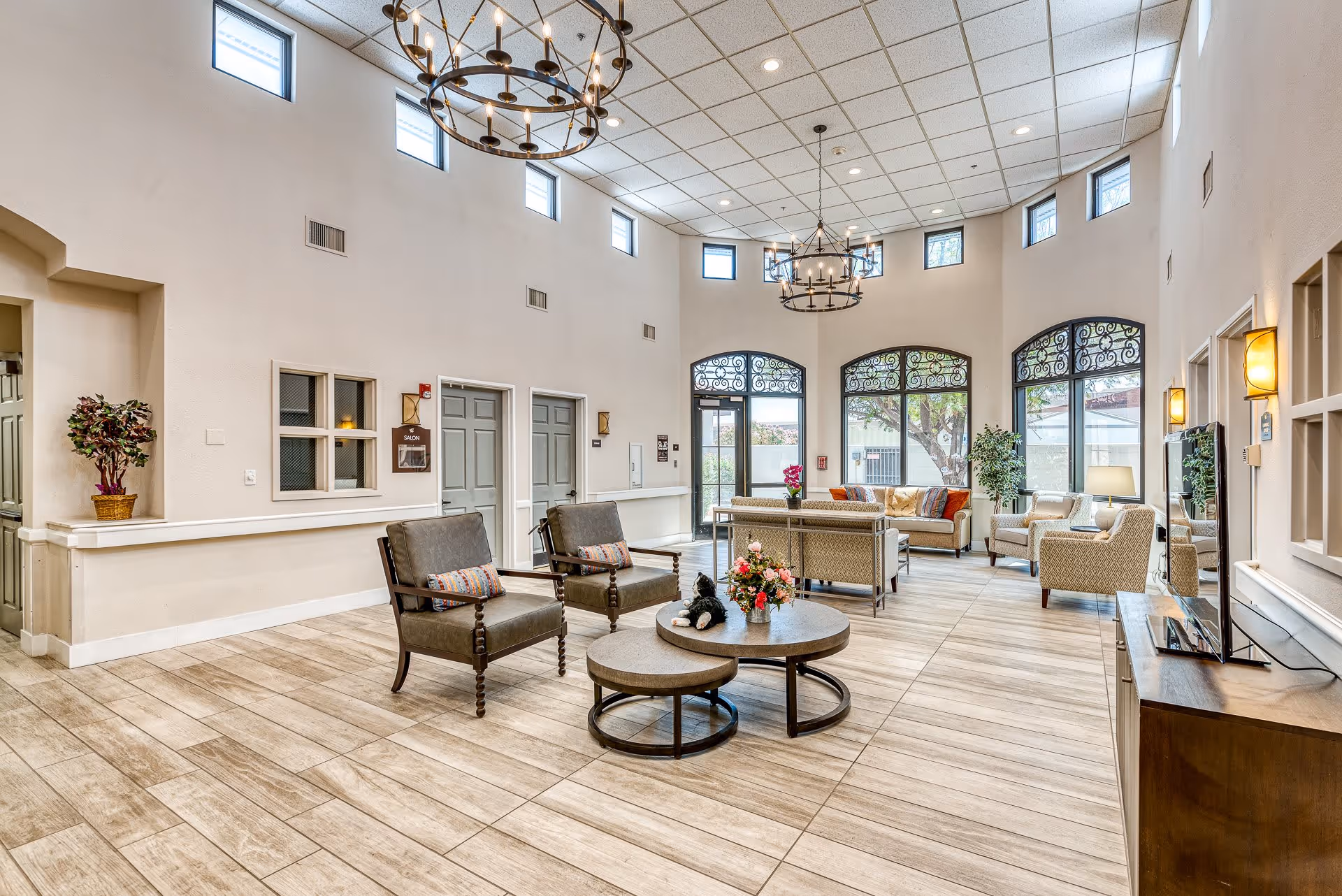 A spacious and well-lit living room area in a senior living facility with high ceilings and multiple small windows near the ceiling. The room features comfortable seating including armchairs and sofas arranged around circular coffee tables with flower arrangements. Large windows with decorative ironwork allow natural light to enter, and there are two large chandeliers hanging from the ceiling. The floor is tiled with a light wood pattern, and there is a TV on a wooden stand to the right.