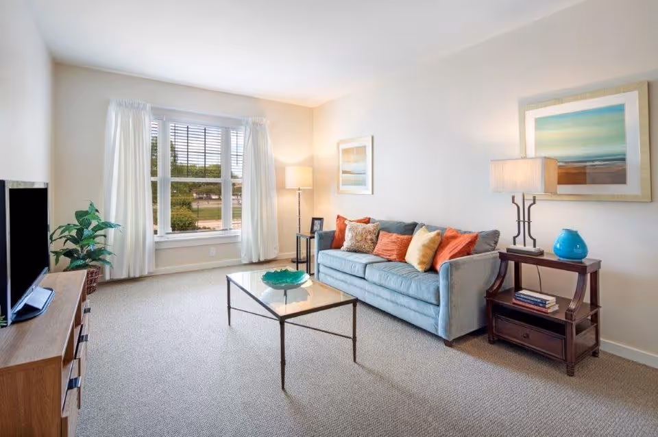 A bright and cozy living room with a large window covered by white curtains, a light blue sofa with multiple colorful cushions, a glass coffee table with a decorative green bowl, a wooden side table with a lamp and a blue vase, a floor lamp, a framed abstract painting on the wall, a wooden TV stand with a flat-screen TV, and a potted plant in the corner.