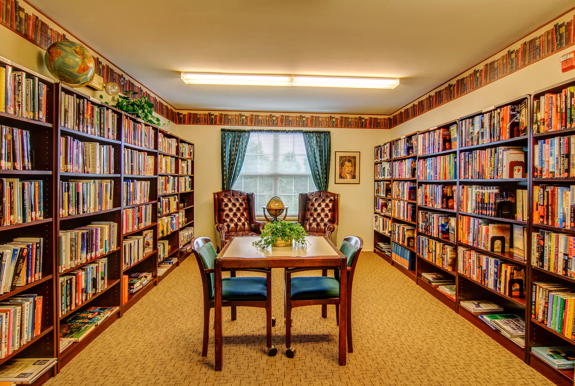 A cozy library room with bookshelves filled with books lining both walls. In the center, there is a wooden table with four chairs around it and a plant on top. At the far end, two brown leather armchairs are positioned in front of a window with green curtains. A globe and framed picture are also visible near the window.