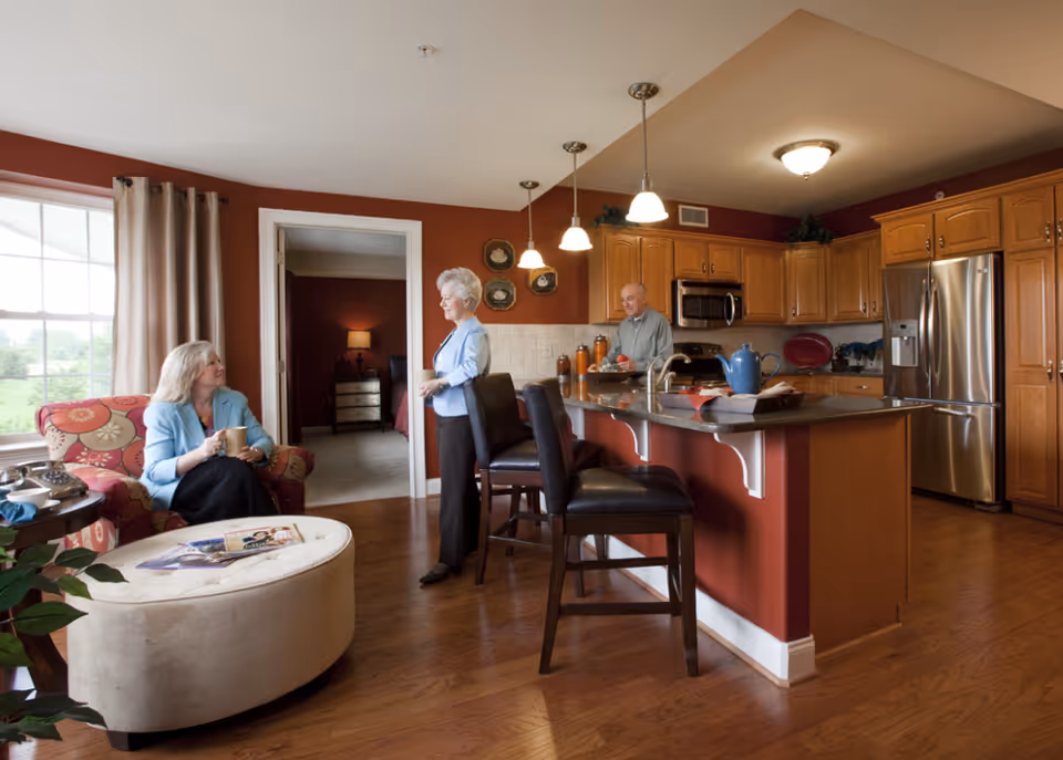 Open-plan kitchen and living area with a kitchen island, seating, and three adults conversing.