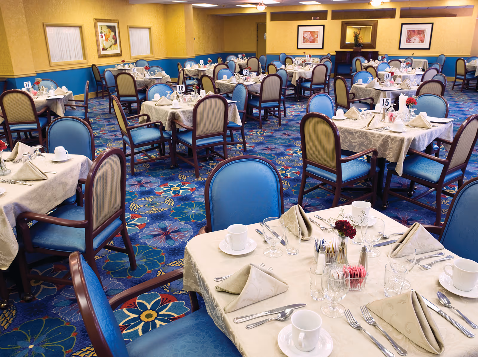 A dining room with multiple tables covered in beige tablecloths, each set with napkins, cups, glasses, and silverware. The chairs have blue cushions and wooden frames. The carpet has a colorful floral pattern, and the walls are painted yellow and blue with framed artwork and a mirror.