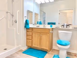 Bright bathroom with a walk-in shower, wooden vanity with a large mirror, and a toilet. The bathroom features turquoise accents including a bath mat, toilet seat cover, and hand towels.
