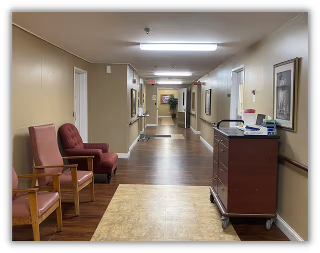 A long hallway in a senior living community with beige walls and wooden flooring. On the left side, there are two chairs, one pink and one red, placed against the wall. On the right side, there is a wooden cart with drawers and various items on top, including a box of tissues and a red container. The hallway is well-lit with ceiling lights and has framed pictures hanging on the walls. At the far end, there is an exit sign and a potted plant near an open door.