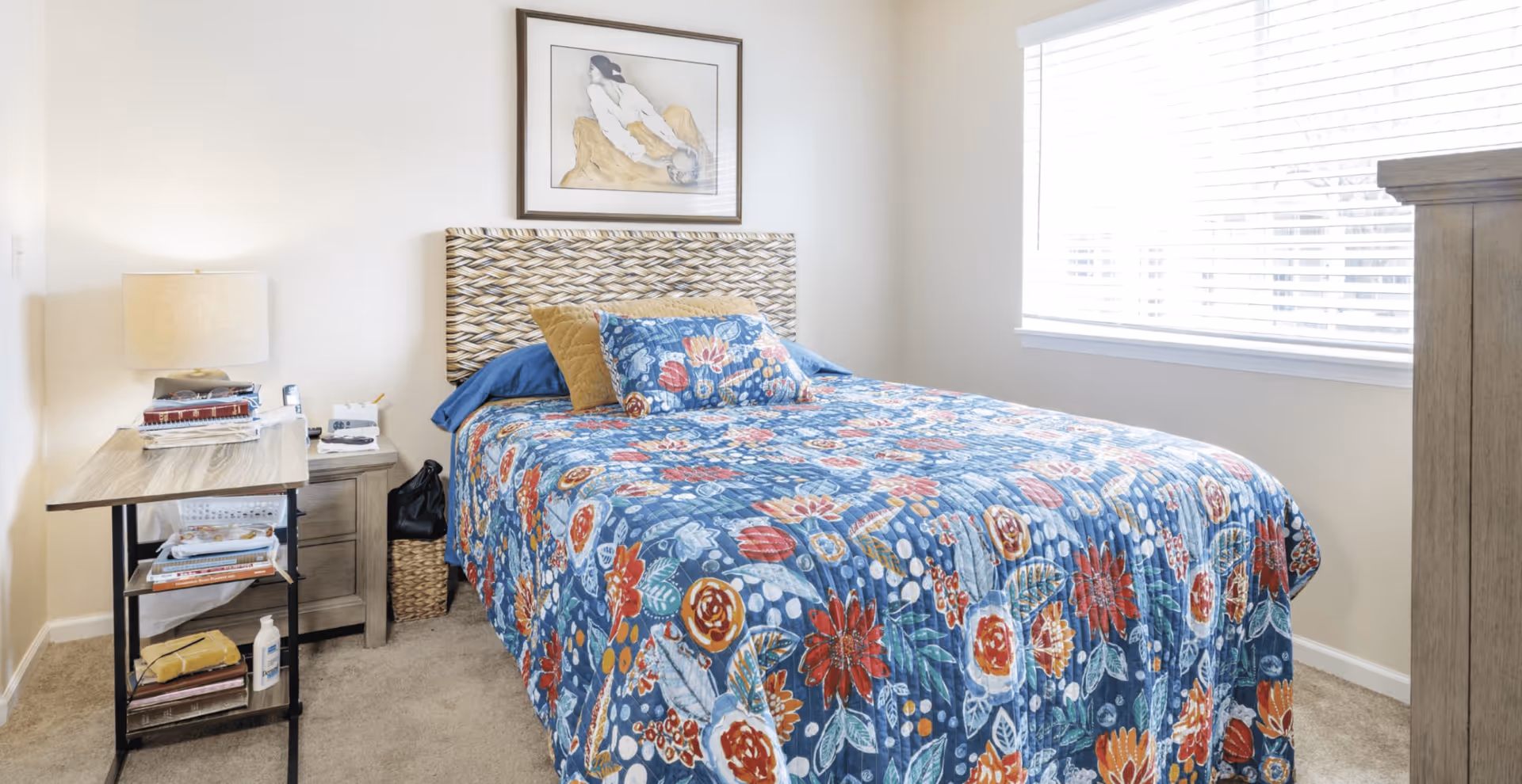 Bright bedroom featuring a bed with a blue floral quilt and woven headboard, a nightstand with a lamp and shelving, and a window with blinds.