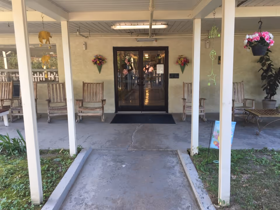 Covered outdoor entrance area with a concrete walkway leading to double glass doors. There are wooden rocking chairs on either side of the entrance, wall-mounted flower decorations, hanging flower pots, and some greenery along the sides of the walkway.