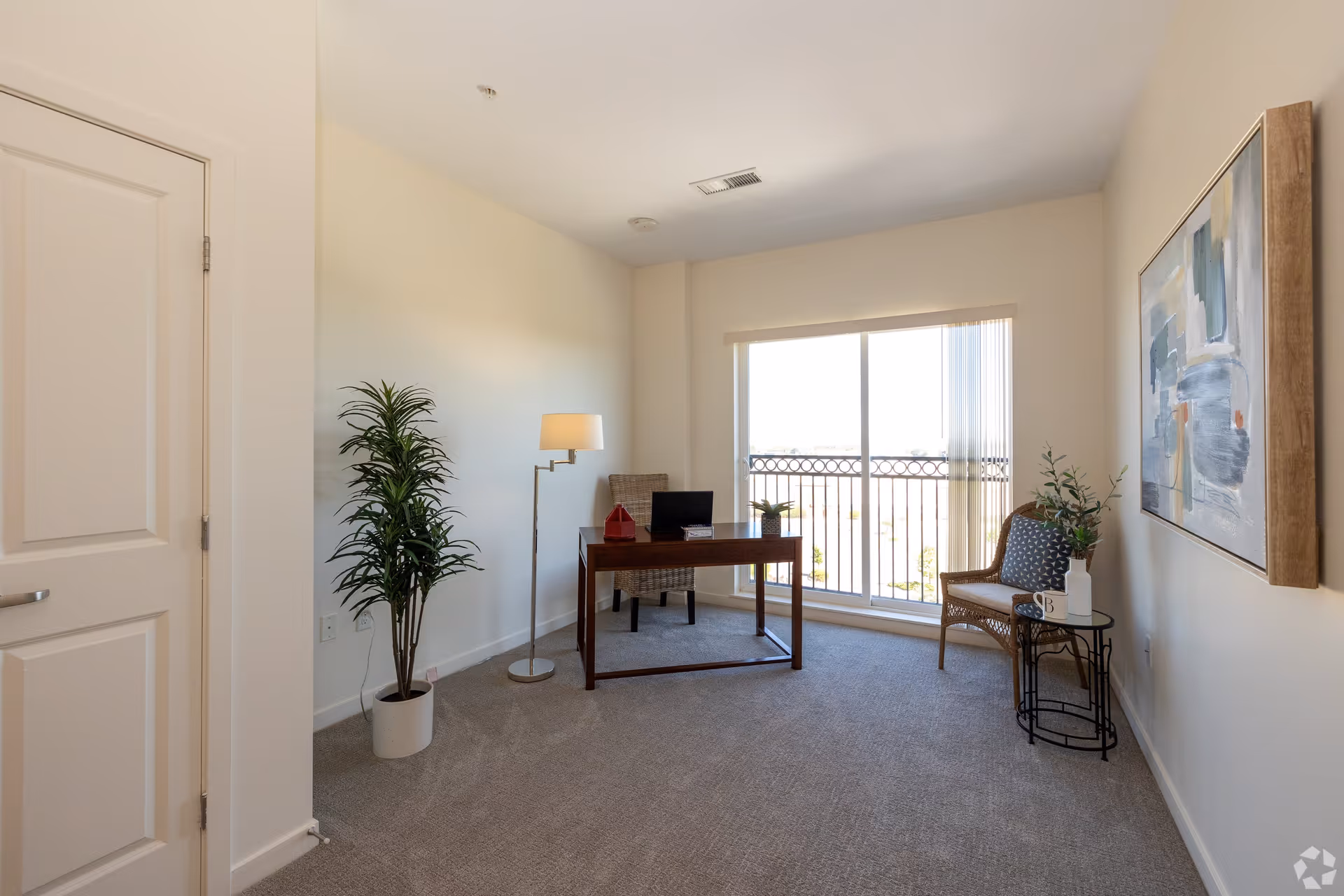 A bright room with beige walls and carpeted floor featuring a wooden desk with a laptop and decorative items, a wicker chair, a small round side table with a plant and a mug, a tall floor lamp, a potted plant, and a large window with vertical blinds letting in natural light.