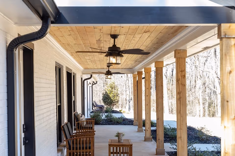 Covered wooden porch with ceiling fans, wooden columns, and chairs overlooking a landscaped outdoor area.