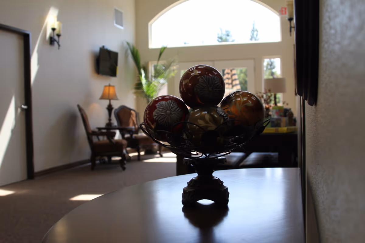 A decorative bowl filled with ornamental balls sits on a wooden table in a softly lit room. In the background, there are two upholstered chairs with a small table and lamp between them, a wall-mounted TV, a large window with an arched top, and a potted plant near the window letting in natural light.