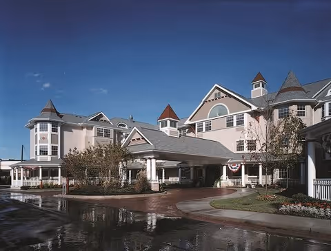 Exterior view of a large, multi-story senior living facility with a covered entrance, multiple peaked roofs, and well-maintained landscaping under a clear blue sky.