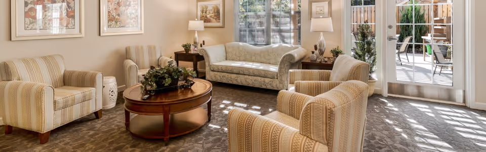 Sunlit communal living room with upholstered armchairs and a sofa arranged around a round wooden coffee table and glass doors to an outdoor patio.