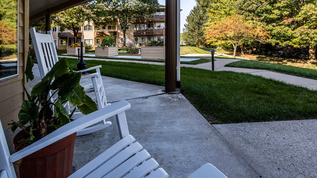 White rocking chairs and a potted plant on a covered patio overlooking a grassy courtyard and apartment building.