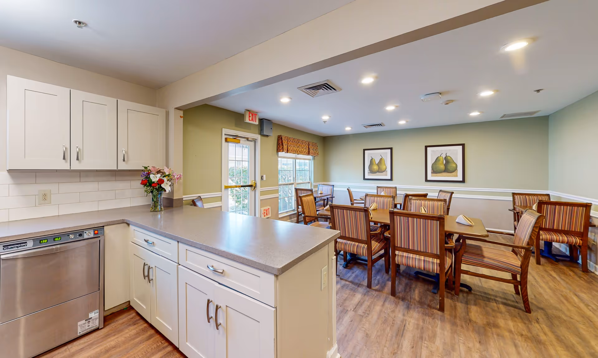Interior view of a dining area in a senior living facility with a kitchen counter in the foreground. The dining area has several wooden tables and chairs with striped upholstery. The walls are painted light green with white trim, and two framed pictures of pears hang on the wall. There is a door and window with a valance letting in natural light.