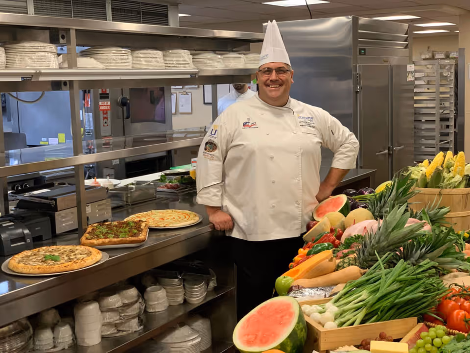 A smiling chef in a commercial kitchen stands behind a counter with pizzas and a large display of fresh fruits and vegetables.