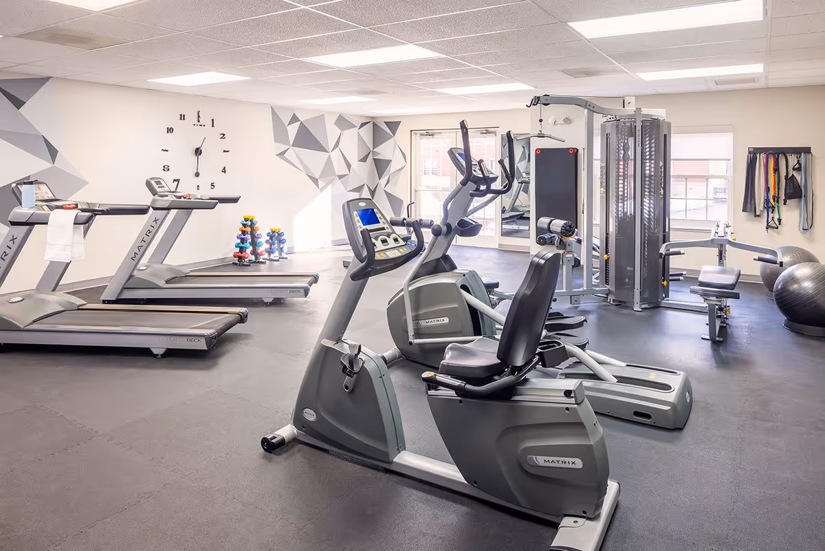 A bright and clean fitness room with exercise equipment including treadmills, stationary bikes, a multi-function weight machine, and free weights. The walls feature geometric patterns and a large clock. There are windows allowing natural light into the room.