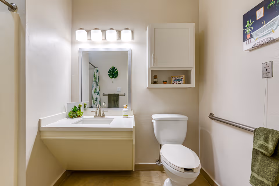 Well-lit bathroom with a vanity and sink under a mirror, a toilet, wall cabinet, and a towel bar.
